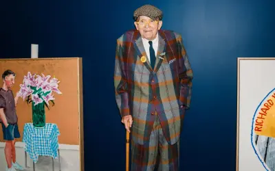 Ein Mann im karierten Anzug, inspiriert von David Hockney Pop Art, steht neben seinen lebendigen Gemälden.