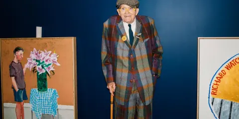 Ein Mann im karierten Anzug, inspiriert von David Hockney Pop Art, steht neben seinen lebendigen Gemälden.