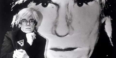 Andy Warhol, Ikone der Pop Art, posiert vor einem riesigen Porträt von sich selbst. Seine Kunstrichtung prägte die moderne Kunst.
