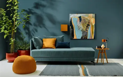 Ein Wohnzimmer mit einem türkisfarbenen Sofa, orangefarbenen Akzenten und einem abstrakten Gemälde. Die Wandfarbe könnte die Farbe des Jahres 2026 sein.