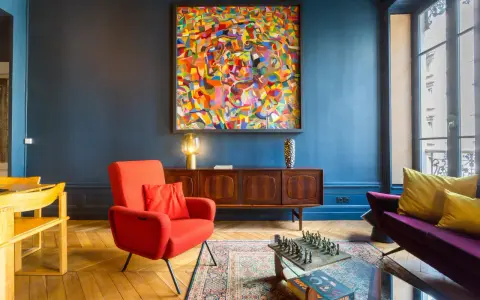 Ein Wohnzimmer mit einem farbenfrohen Gemälde im art déco zeitraum, rotem Sessel, Holzsideboard und einem Schachspiel auf dem Couchtisch.