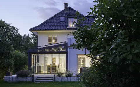 Moderner Anbau Wintergarten mit Glasfront und Beleuchtung erweitert ein Haus. Der Garten ist abends beleuchtet.