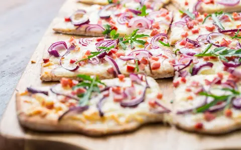 Flammkuchen-Ideen: Dünner Teig mit Käse, roten Zwiebeln, Speck und Rucola belegt. Perfekt für den Sommer!