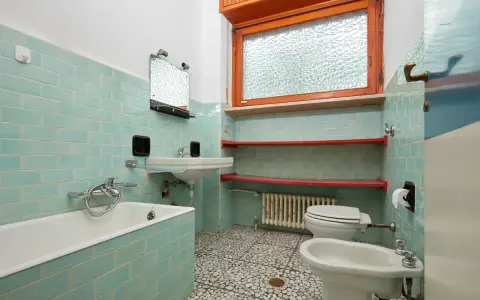 Bad renovieren ohne fliesen abschlagen: hellblau geflieste Wände, Badewanne, Waschbecken, Toilette, Bidet und Fenster.