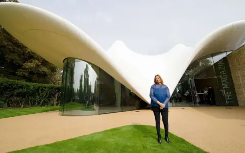 Zaha Hadid steht vor einem ihrer ikonischen zaha hadid bauwerke, einem geschwungenen, weißen Dach mit Glasfront.