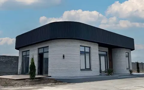 Ein modernes 3d druck haus mit runden Ecken und einem dunklen Dach steht unter einem blauen Himmel mit Wolken.