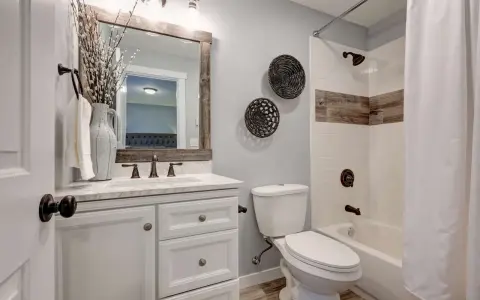 Ein kleines Bad gestalten 4qm: Weißer Waschtisch mit Marmorplatte, Spiegel mit Holzrahmen, Toilette, Duschbadewanne mit weißem Vorhang und rustikalen Akzenten.