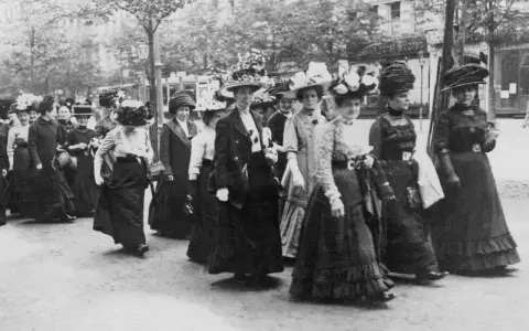 Frauen in langen Kleidern und Hüten marschieren entschlossen. Ein historisches Bild der feministischen Bewegung.