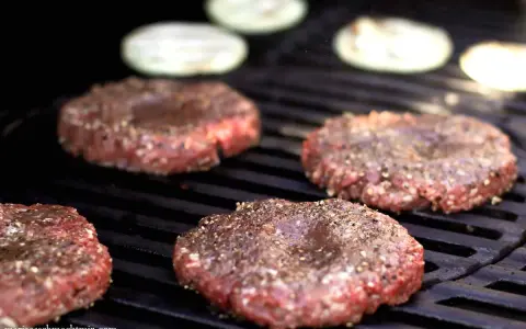 Vier frisch geformte Patties, gewürzt mit Pfeffer, brutzeln auf dem Grill. Perfekt, um Patties selber machen.