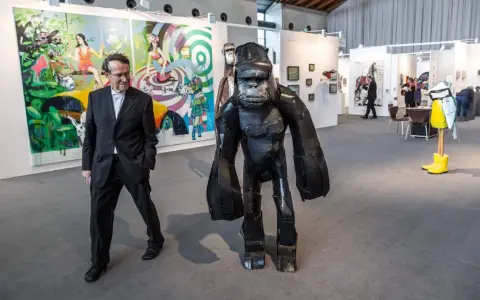 Mann in Anzug neben Gorilla-Skulptur auf Kunstmesse. Bunte Gemälde im Hintergrund zeigen aktuelle Kunst.