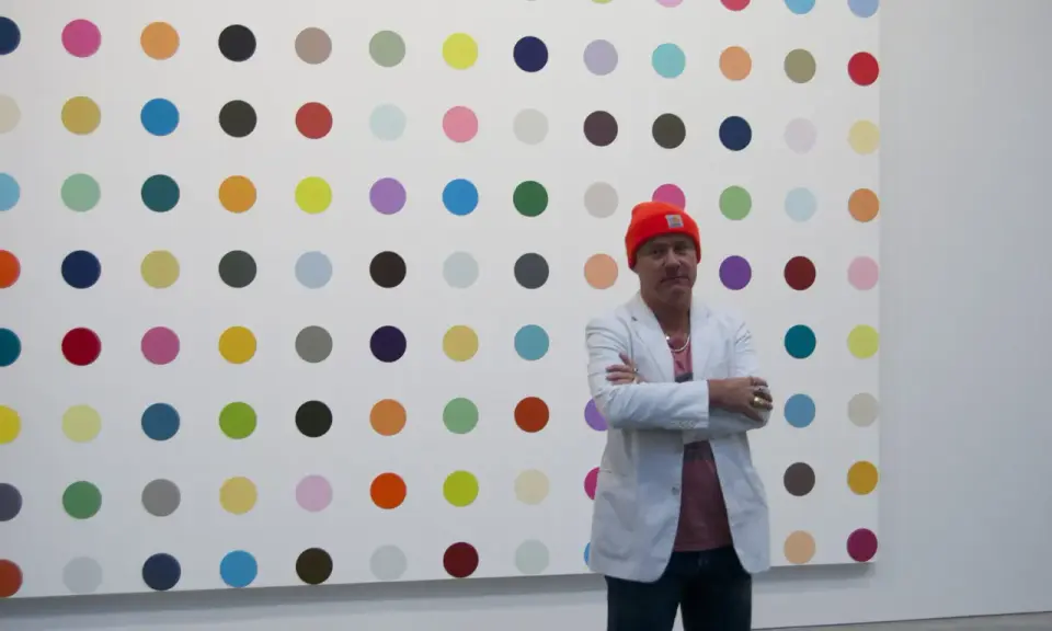 Damien Hirst steht vor seiner berühmten Punkt-Malerei, gekleidet in eine orangefarbene Mütze und einen weißen Blazer.