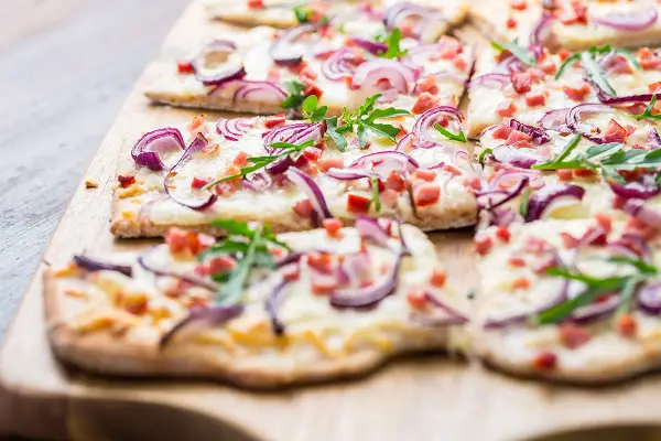 Flammkuchen-Ideen: Dünner Teig mit Käse, roten Zwiebeln, Speck und Rucola belegt. Perfekt für den Sommer!