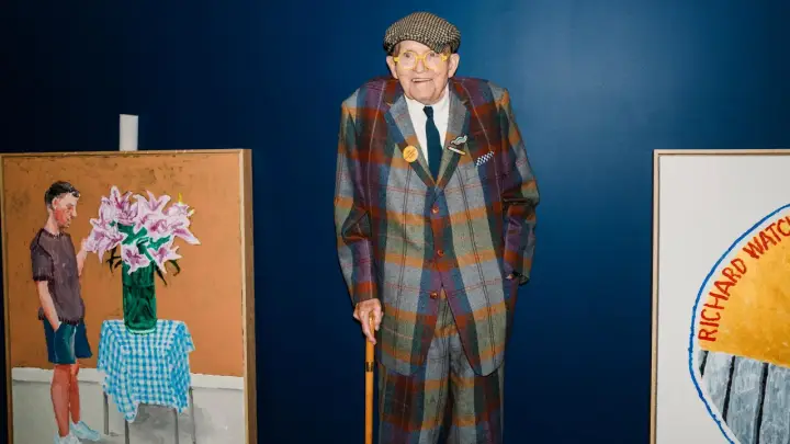 Ein Mann im karierten Anzug, inspiriert von David Hockney Pop Art, steht neben seinen lebendigen Gemälden.
