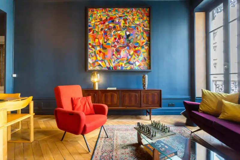 Ein Wohnzimmer mit einem farbenfrohen Gemälde im art déco zeitraum, rotem Sessel, Holzsideboard und einem Schachspiel auf dem Couchtisch.